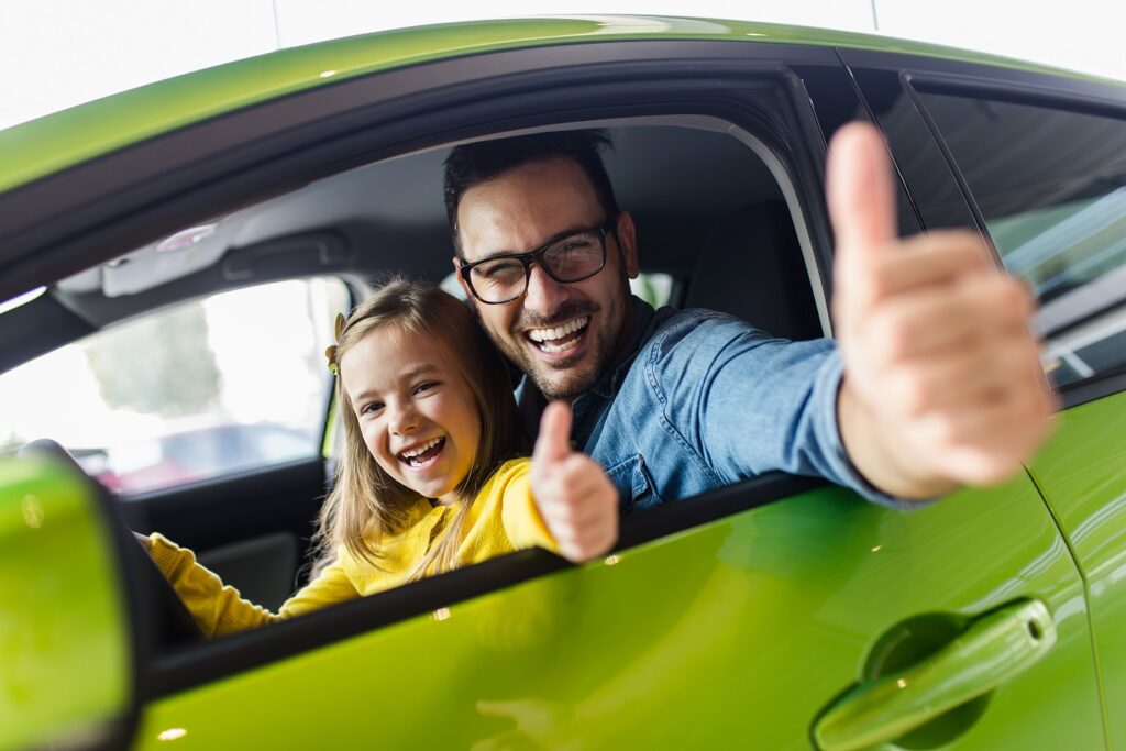Father and daughter buying a new car at the car showroom and giving thumbs up to the camera.